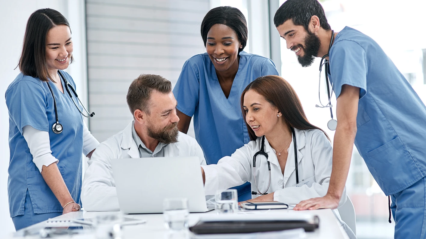 Medical professionals collaborate around a laptop, discussing strategies to improve care delivery and patient outcomes.