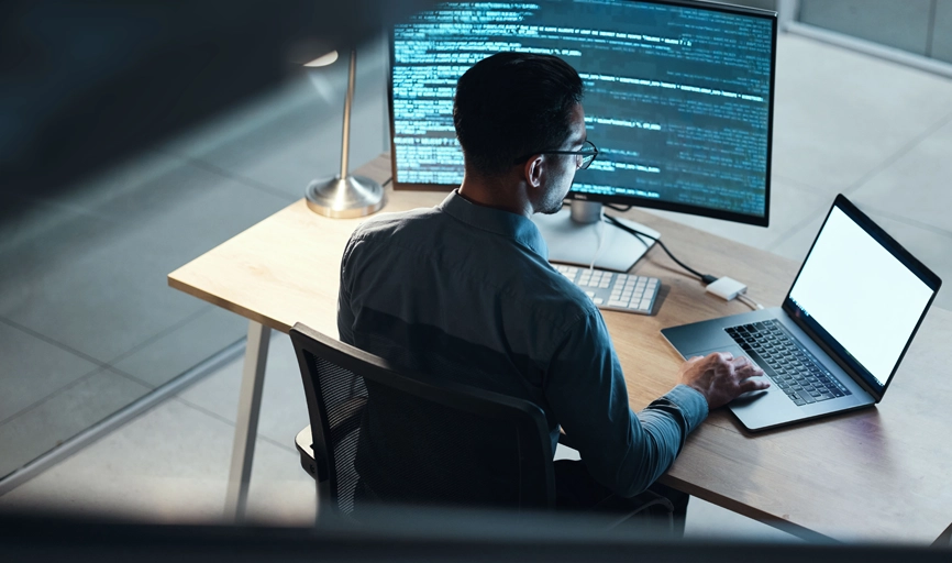 Black male programmer seen from behind using his technical expertise on a laptop connected to a large monitor