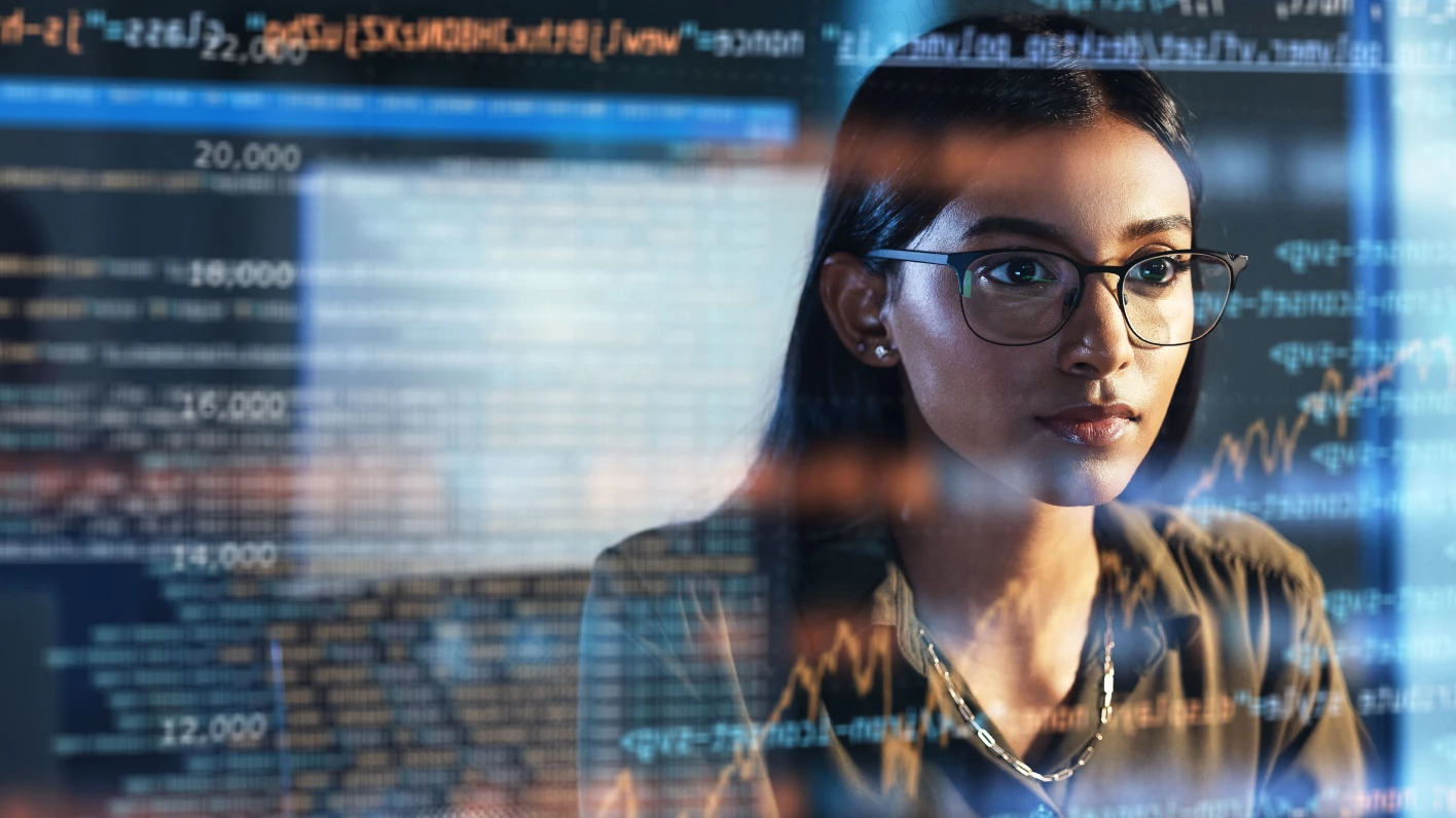 Woman in glasses stares intently at a screen, seen in reflection behind her. Overlay of soft-focus coding covers the screen.
