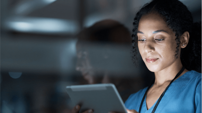 Woman in blue smock is peering at a tablet in a darkened room, confident that Information security risk management is in place.