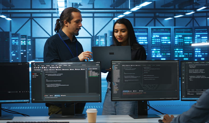 Two Scrum Masters: man with dark hair, ponytail and Indian woman conferring in a dimly lit room with servers in background