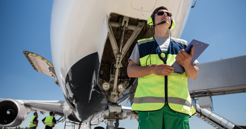 Wide angle of man in hard hat & safety vest in front of jet on runway with a tablet utilizing new data platform.