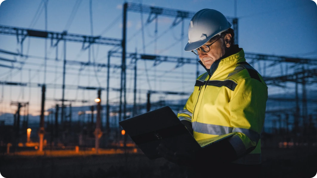 Man in hard hat and yellow safety jacket stands in front of a bank of servers with a clipboard.