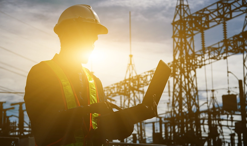 Silhouette of a man in hard hat against sunset. He holds a laptop symbolizing new data ecosystem. Background is a power station