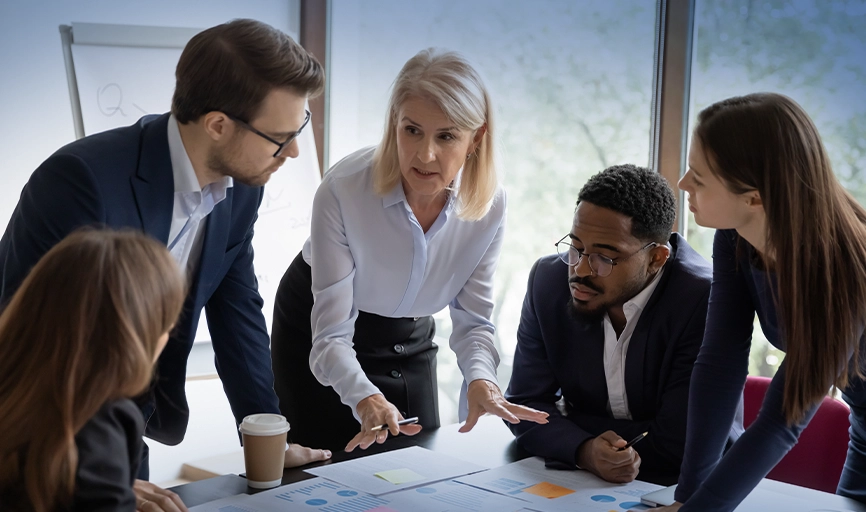 Content Manager Blonde white woman, a pharma software provider in business attire, is explaining something to four colleagues around a conference table.